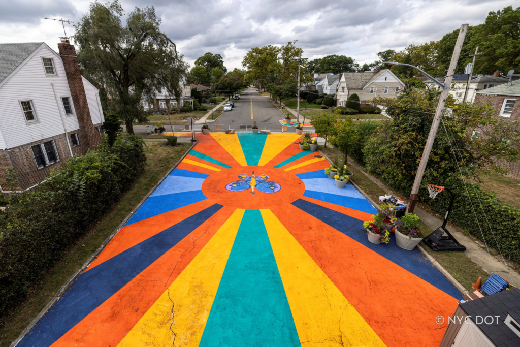 A vibrant asphalt art mural, titled Magnify! Legacy! Triumph!, on the new pedestrian plaza at the Murdock Avenue Open Street in Addisleigh Park, Queens. Image Credit: NYC DOT