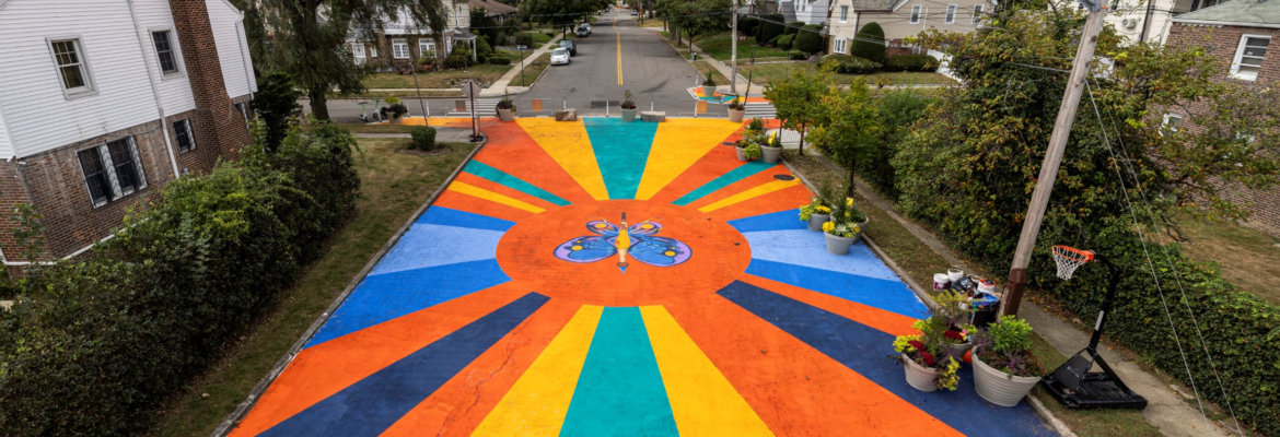 A vibrant asphalt art mural, titled Magnify! Legacy! Triumph!, on the new pedestrian plaza at the Murdock Avenue Open Street in Addisleigh Park, Queens. Image Credit: NYC DOT